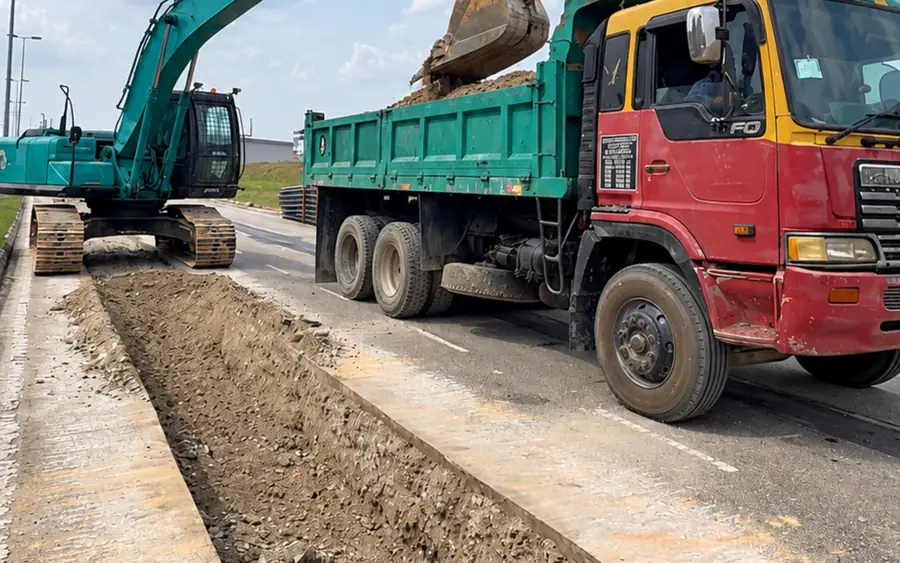 Services Green City Machinery Kobelco excavator trenching along a road reserve and loading a lorry tipper for underground services in Johor